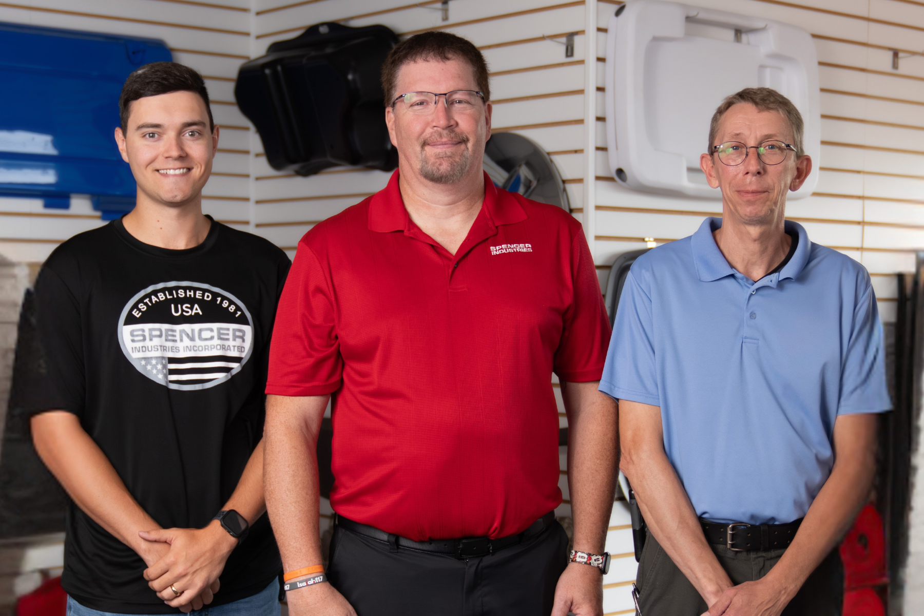 Three men stand side by side in a hardware store or workshop, smiling at the camera. The left man wears a black T-shirt, the middle man in a red polo, and the right man in a light blue polo.