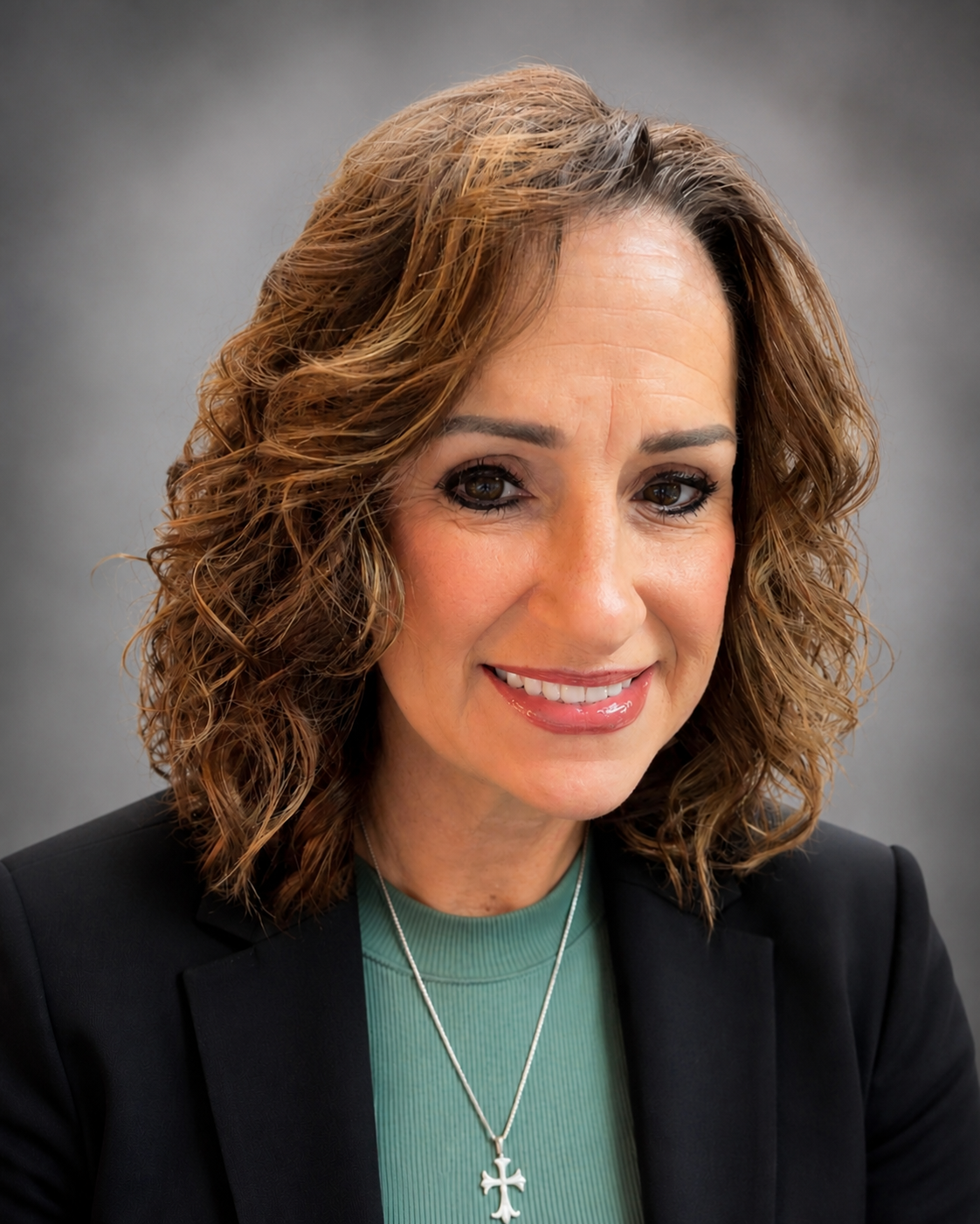 Smiling woman with shoulder-length curly brown hair, wearing a black blazer over a green top and a silver cross necklace, in a neutral studio background.